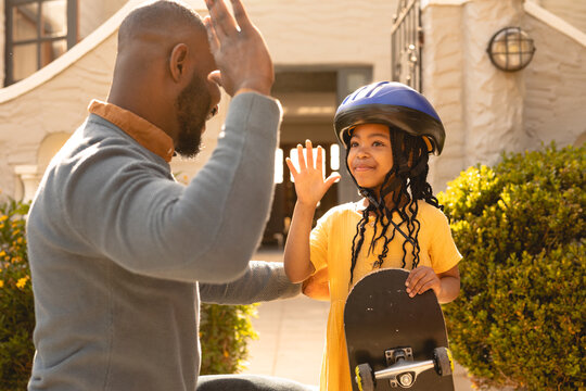 Smiling african american girl wearing helmet holding skateboard giving high-five to father at yard