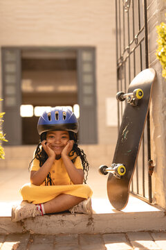 Portrait of smiling african american girl wearing helmet sitting cross-legged by skateboard at gate