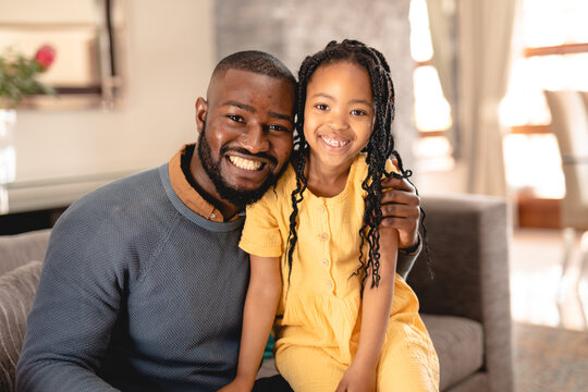 Portrait of smiling african american man sitting with arm around daughter on sofa in living room