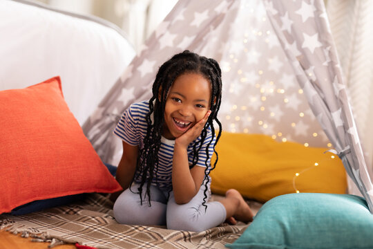 Portrait of cheerful cute african american girl kneeling on bed with tent and cushions at home