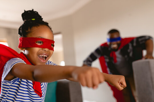 Happy african american girl wearing superhero costume flying in front of father playing at home