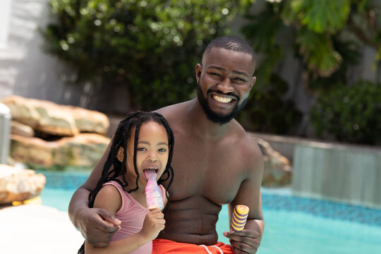 Portrait of happy african american father and daughter eating ice cream and pops at poolside - Powered by Adobe
