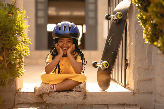 Portrait of cheerful african american girl wearing helmet sitting cross-legged by skateboard at gate