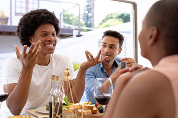 Happy young biracial man with afro hairstyle talking to friends sitting together at dining table