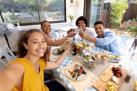 Smiling young biracial woman taking selfie while toasting drinks at dining table