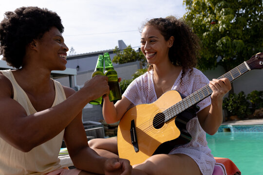 Happy biracial woman with guitar toasting beer bottle with man at poolside on sunny day