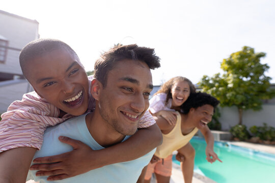 Young biracial men giving piggyback rides to cheerful women at poolside on sunny day - Powered by Adobe