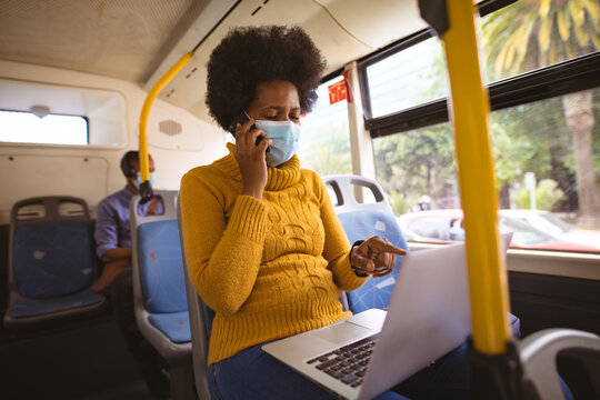 African american mid adult businesswoman with mask talking on call while using laptop in bus