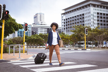 African american mid adult businesswoman with wheel luggage looking away while crossing crosswalk