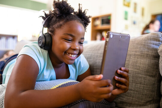 Smiling african american girl with digital tablet wearing headphones while lying on sofa at home