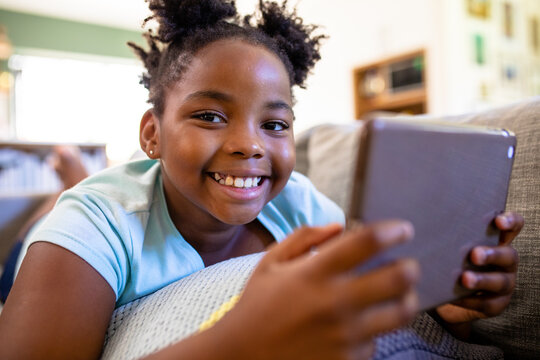 Portrait of smiling african american girl with digital tablet lying on sofa at home