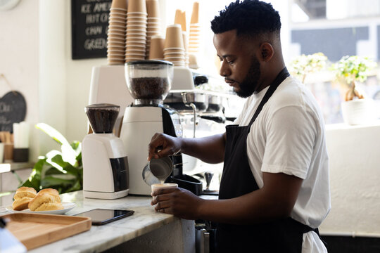 Confident young african american barista adding milk in coffee cup while working at cafe