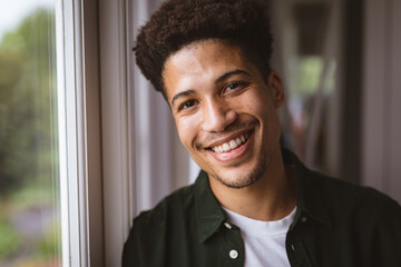 Portrait of cheerful biracial young man in casuals by window at home