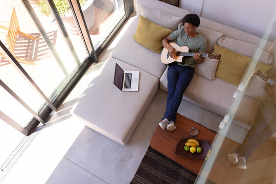 African American Young Man Playing Guitar While Sitting With Laptop And Feet Up On Table At Home