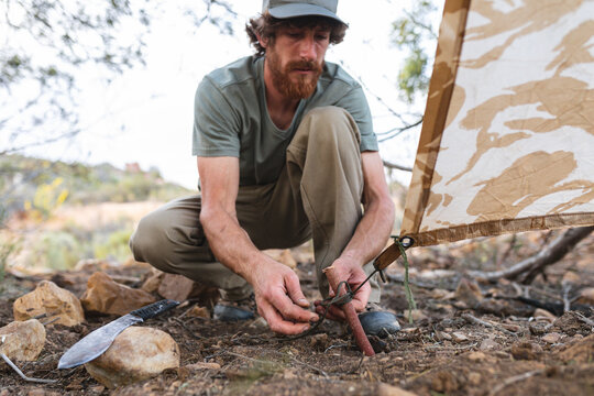 Full length of young bearded male caucasian adventurer pitching tent while crouching in forest