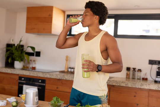 African american young man drinking smoothie while standing in kitchen at home - Powered by Adobe