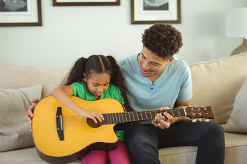 Happy biracial father teaching daughter to play guitar while sitting together on sofa in living room