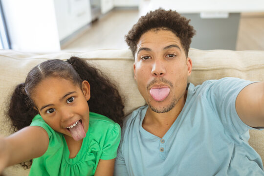 Playful biracial father and daughter taking selfie while sticking out tongue at home