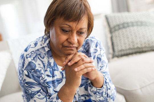 Depressed african american senior woman sitting with hands clasped in living room at home