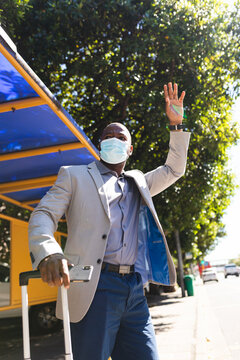 African american businessman wearing a face mask with suitcase hailing a taxi on the go to office