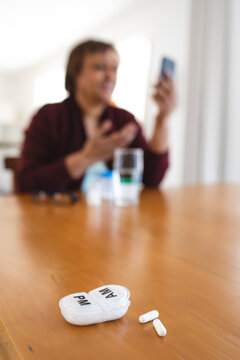 Pill box and capsules on table while woman taking online doctor consultation on smart phone at home