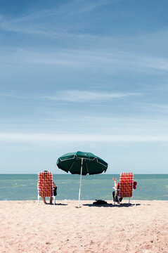 Two women sitting on deck chairs under an umbrella facing the sea