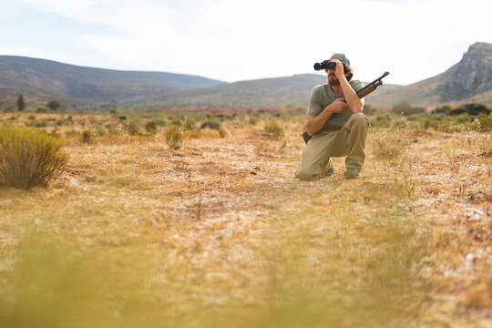 Full length of young caucasian male hunter crouching with rifle while looking through binoculars