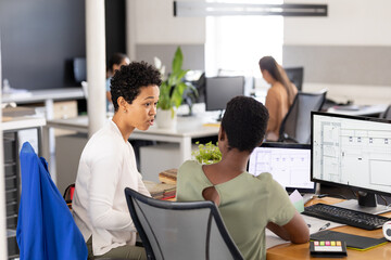 Female architects with blueprints on computer discussing at desk in creative office
