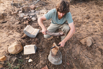 High angle view of young male caucasian survivalist cutting firewood with knife in forest