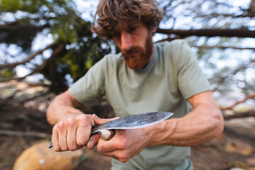 Dedicated bearded young caucasian male survivalist sharpening stick with knife in forest