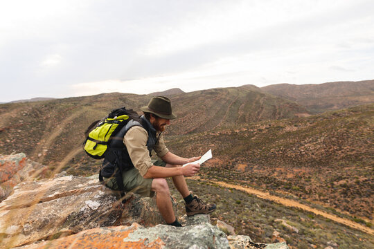 Side view of young male caucasian adventurer looking at map while sitting on rock at mountain peak
