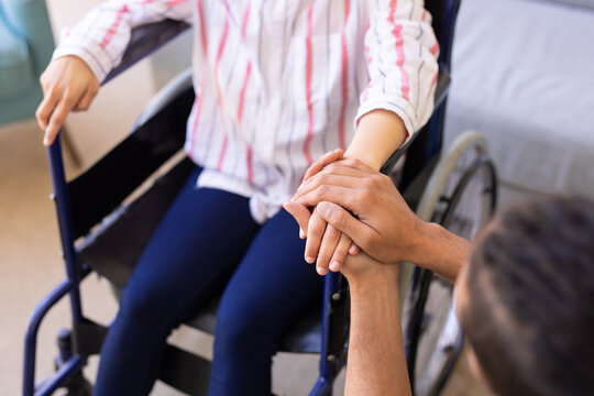 Mid section of a man holding hands of his disabled wife sitting on wheelchair at home