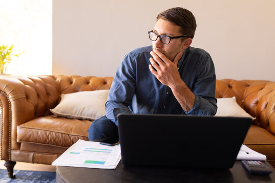 Thoughtful caucasian man using laptop and calculating finances at home - Powered by Adobe