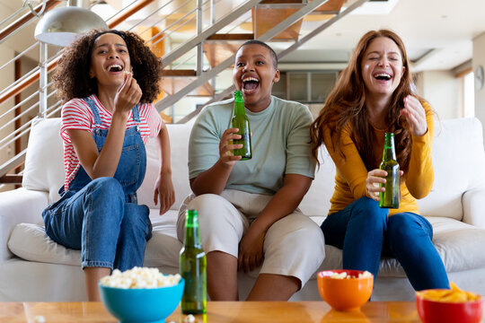 Multiracial female friends with beer and popcorn laughing while watching tv at home - Powered by Adobe