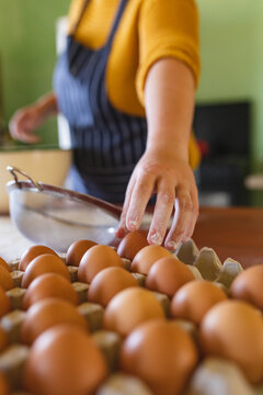 Midsection of woman with flour on fingers picking brown egg from carton in kitchen
