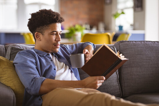Young biracial handsome man holding coffee mug reading book while relaxing on sofa at home