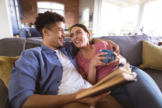 Happy loving biracial couple looking at each other while relaxing with coffee and book at home