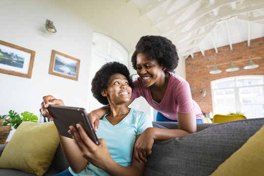 Happy mother looking at teenage daughter using digital tablet on sofa at home