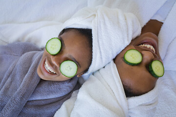 Mother and daughter in bathrobes smiling while relaxing with cucumber slices on eyes at spa
