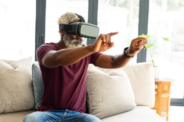 African american senior man gesturing while watching virtual reality through headset sitting on sofa