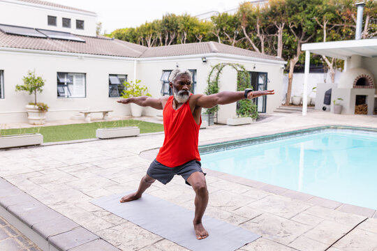 Senior african american man practicing warrior pose yoga on mat at poolside - Powered by Adobe