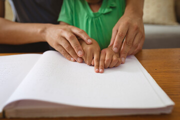 Midsection of biracial father helping blind daughter studying while reading book with braille