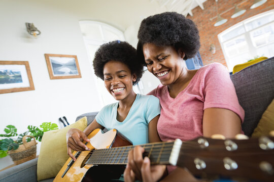 Cheerful mother with teenage daughter practicing guitar in living room at home
