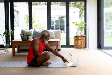 Senior african american man using laptop while sitting on yoga mat in living room at home