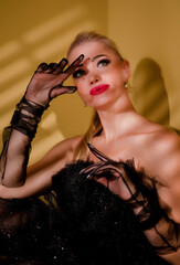 A beautiful blonde in a lush shiny black dress on a yellow background wearing gloves.