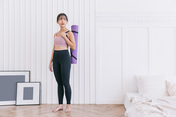 Asian woman wearing sportswear and yoga pants carrying a rolled-up yoga mat standing in a bedroom with sunlight from the window. Working out at home, weight loss concepts. Image with copy space.