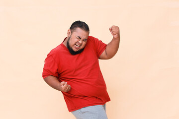 Atrractive young fat Asian man raising his fists with smiling delighted face, yes gesture,...