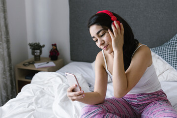 Young latin woman listening music with headphones on bed at home in Mexico Latin America	