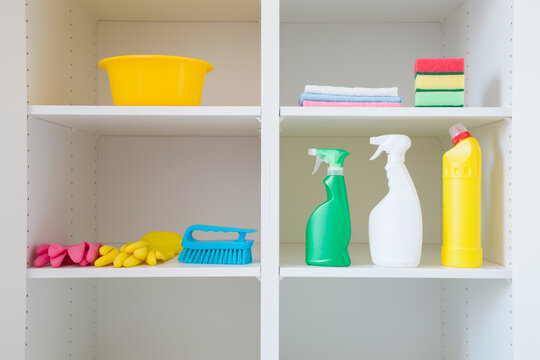 Bowl, brush, detergent bottles, rags, rubber gloves and sponges on white shelves inside opened wardrobe. Closeup. Set of colorful products for house cleaning. Front view. Home neatness.