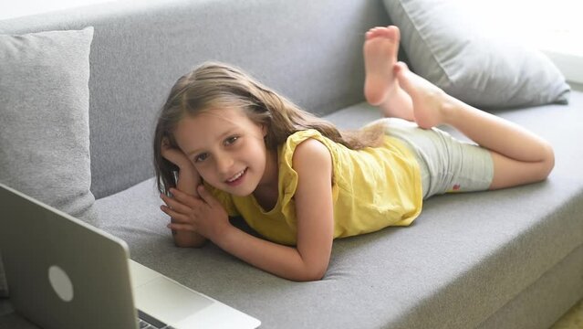 Cute child girl using laptop, lies on couch, little smart kid typing on computer chatting with friend, talk online, study at home, playing and watch cartoons on gadget.